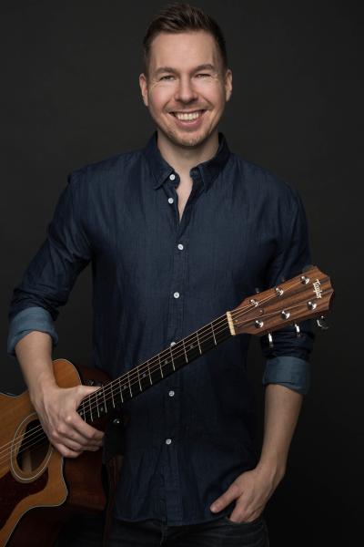 Komiker Dan Andersen står og smiler med front mod kameraet med en guitar i højre hånd